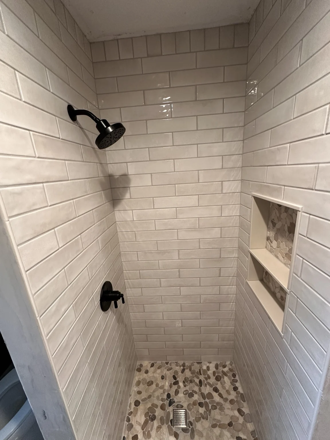 White subway tile shower with black fixtures, pebble floor and recessed shelving